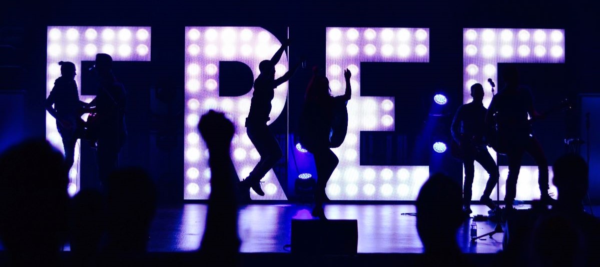 Band playing in front of large lit up letters spelling FREE, photo by William White on Unsplash